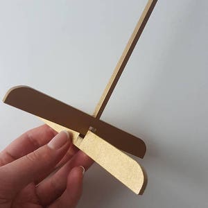 Op de afbeelding: Een gouden houten ezelstandaard met een lange, dunne verticale steun en een kruisvormige basis. De basis is gemaakt van twee stukken hout die in een rechte hoek aan elkaar zijn bevestigd.