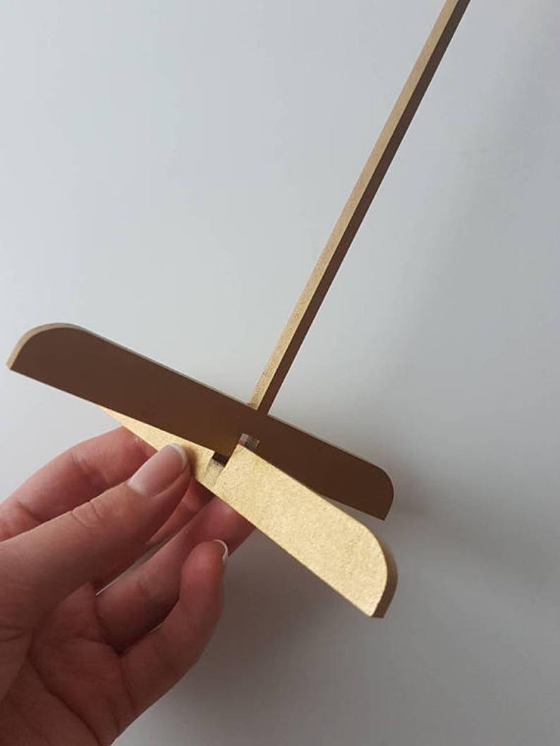 Op de afbeelding: Een gouden houten ezelstandaard met een lange, dunne verticale steun en een kruisvormige basis. De basis is gemaakt van twee stukken hout die in een rechte hoek aan elkaar zijn bevestigd.