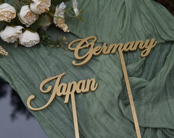 May include: Two gold cake toppers, one reading "Germany" and the other "Japan", in a cursive script. They rest on a sage green fabric, with white flowers in the upper left.