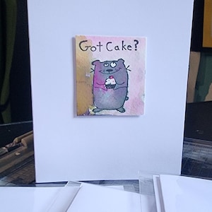 Könnte beinhalten: Weiße Grußkarte mit einer farbenfrohen Illustration eines Cartoon-Bären, der einen Cupcake hält. Der Text "Got Cake?" steht über dem Bären. Mehrere andere Karten sind im Vordergrund zu sehen.