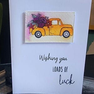 Könnte beinhalten: Weiße Grußkarte mit dem handgeschriebenen Text "Wishing you loads of luck". Die Karte zeigt ein Aquarellgemälde eines gelben Pick-ups mit lila und roten Blumen.