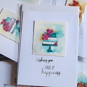 Könnte beinhalten: Aquarell-Grußkarten mit einem türkisfarbenen Kuchen mit rosa und orangefarbenen Blumen. Die Karten tragen den Text "Wishing you loads of happiness". Die Karten sind in weißen Umschlägen.