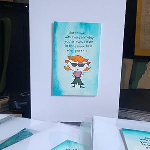 Könnte beinhalten: Humorvolle Grußkarte mit Cartoon-Illustration. Die Karte zeigt eine Zeichentrickfigur mit orangefarbenem Haar und Sonnenbrille. Der Text auf der Karte lautet: „Denk nur, mit jedem Geburtstag kommst du deinen Eltern immer näher.“