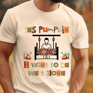 May include: Cream-colored t-shirt with the text "Yes Pumpkin" and "I want to be left alone" in various colors and fonts. The shirt features a graphic of a person on a swing, pumpkins, and watering cans.