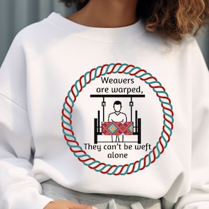 Peut inclure: Sweat-shirt blanc avec un motif circulaire représentant un tisserand et le texte "Weavers are warped, They can't be weft alone." Le motif est encadré par un motif de corde rouge, turquoise et blanche. Le sweat-shirt est de style décontracté et confortable.