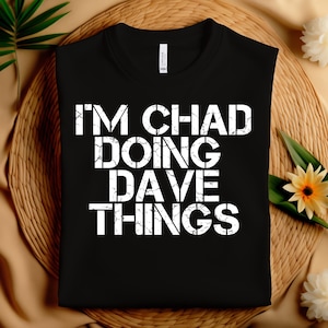May include: Black t-shirt with the white text "I'M CHAD DOING DAVE THINGS". The shirt is folded and placed on a woven surface with floral accents. The text is in a bold, cracked font.