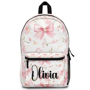 May include: White backpack with a pink bow and floral pattern. The backpack has a black zipper, a black handle, and a black mesh side pocket. The name "Olivia" is printed in black script on the front pocket.