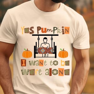 May include: White t-shirt with the text "Yes Pumpkin, I want to be left alone." The design includes a person on a swing, two orange pumpkins, and text in various colors and fonts. The shirt is a casual wear item.