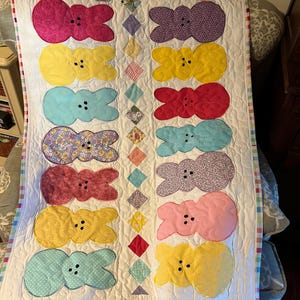 May include: A white quilt with a patchwork design featuring a row of colorful Peeps marshmallow candy shaped appliques. The quilt is edged with a pink and white checkered border.