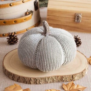 Peut inclure: Une citrouille en tricot gris sur une tranche de bois. La citrouille a une texture côtelée et un sommet légèrement pointu.