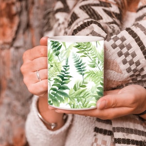 May include: White ceramic mug with a green fern pattern. The mug is being held by a person.