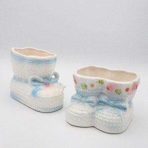 Peut inclure: Deux pots de fleurs en forme de chaussures de bébé en céramique. Le pot de gauche est une seule chaussure avec un ruban bleu et un nœud. Le pot de droite est une paire de chaussures avec un ruban bleu et un nœud. Les deux pots ont un fond blanc avec des accents bleus et roses.
