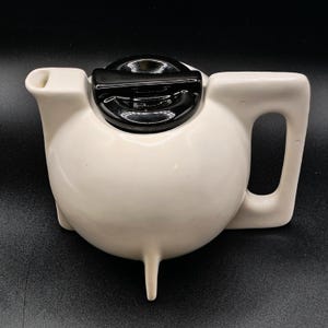 May include: A white ceramic teapot with a black lid and handle. The teapot has a rounded body, a spout, and a handle. The lid is rectangular and sits on top of the teapot. The teapot has a single leg.