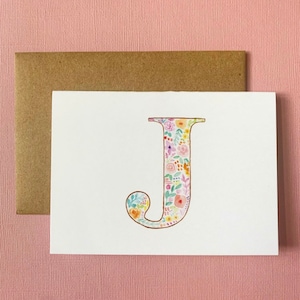 May include: A white card with a floral letter J. The letter is filled with a colorful floral pattern and outlined in gold.