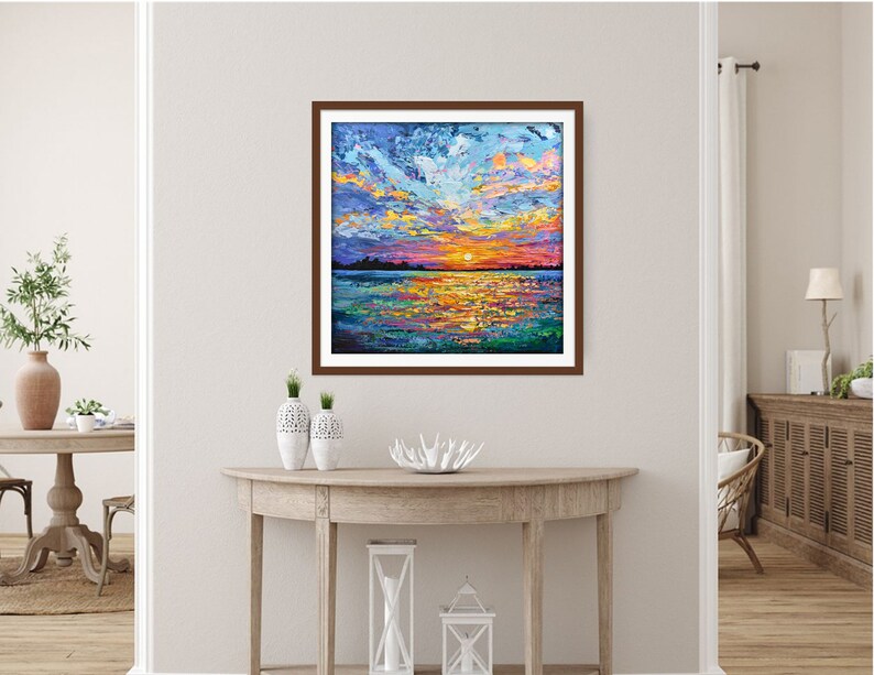 May include: A framed painting of a colorful sunset over a lake. The sky is a mix of blue, purple, and orange, with clouds reflecting in the water. The sun is setting behind the horizon, casting a warm glow on the scene.