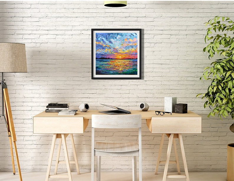 May include: A framed landscape painting depicts a vibrant sunset over water. The artwork features bold brushstrokes of orange, yellow, blue, and purple, creating a textured and colorful scene. The painting is displayed on a white brick wall.