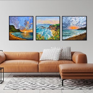 May include: Three framed landscape paintings depicting ocean scenes with vibrant colors. Each artwork captures a sunset or sunrise over the water, with sandy beaches and rocky cliffs. The paintings are displayed above a brown leather sofa.