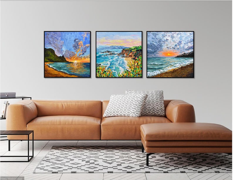 May include: Three framed landscape paintings depicting ocean scenes with vibrant colors. Each artwork captures a sunset or sunrise over the water, with sandy beaches and rocky cliffs. The paintings are displayed above a brown leather sofa.