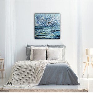 K&ouml;nnte beinhalten: Abstraktes Gem&auml;lde einer Meereslandschaft in Blau-, Wei&szlig;- und Goldt&ouml;nen mit strukturiertem, pastosem Finish. Das Kunstwerk h&auml;ngt &uuml;ber einem Bett mit einer grauen Bettdecke und einer cremefarbenen Strickdecke. Es erinnert an Meer und Himmel.
