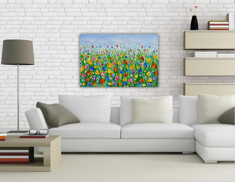 May include: A painting of a field of colorful flowers with a blue sky in the background. The flowers are painted in a variety of shades of red, orange, yellow, pink, blue, and purple. The painting is framed and hanging on a white brick wall.