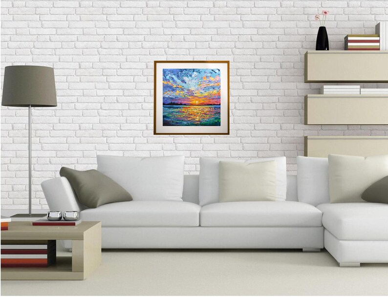 May include: A white couch with gray and cream pillows in front of a white brick wall. A framed painting of a colorful sunset over a body of water hangs on the wall.