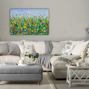 Puede incluir: Una vibrante pintura al &oacute;leo texturizada representa un campo de flores coloridas bajo un cielo azul claro. La obra de arte presenta una variedad de tonos brillantes, incluyendo rojo, amarillo, azul y morado, creando una escena alegre. La pintura se exhibe sobre un sof&aacute; blanco.