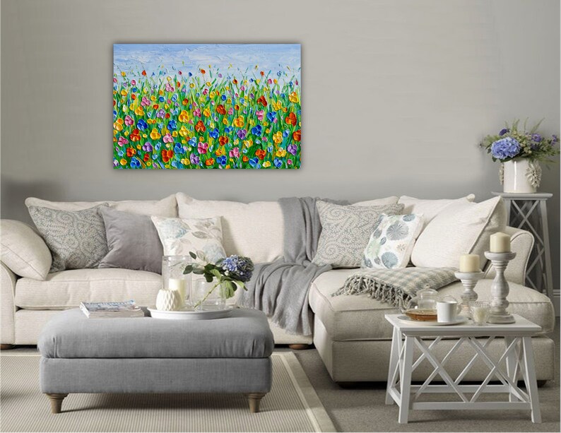 Puede incluir: Una vibrante pintura al &oacute;leo texturizada representa un campo de flores coloridas bajo un cielo azul claro. La obra de arte presenta una variedad de tonos brillantes, incluyendo rojo, amarillo, azul y morado, creando una escena alegre. La pintura se exhibe sobre un sof&aacute; blanco.