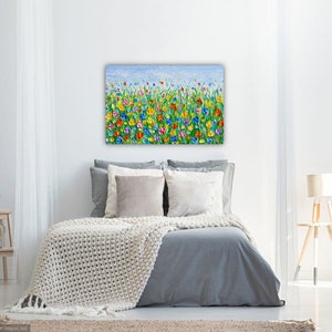 Puede incluir: Una colorida pintura floral sobre lienzo, que presenta un campo de flores vibrantes en tonos de rojo, amarillo, azul y morado contra un cielo azul claro. La obra de arte se exhibe sobre una cama con ropa de cama gris y una manta color crema.