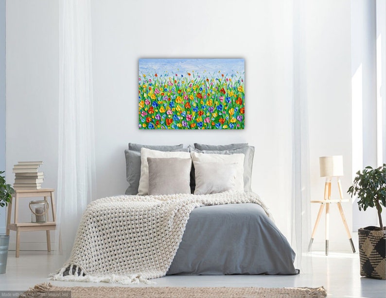 Puede incluir: Una colorida pintura floral sobre lienzo, que presenta un campo de flores vibrantes en tonos de rojo, amarillo, azul y morado contra un cielo azul claro. La obra de arte se exhibe sobre una cama con ropa de cama gris y una manta color crema.
