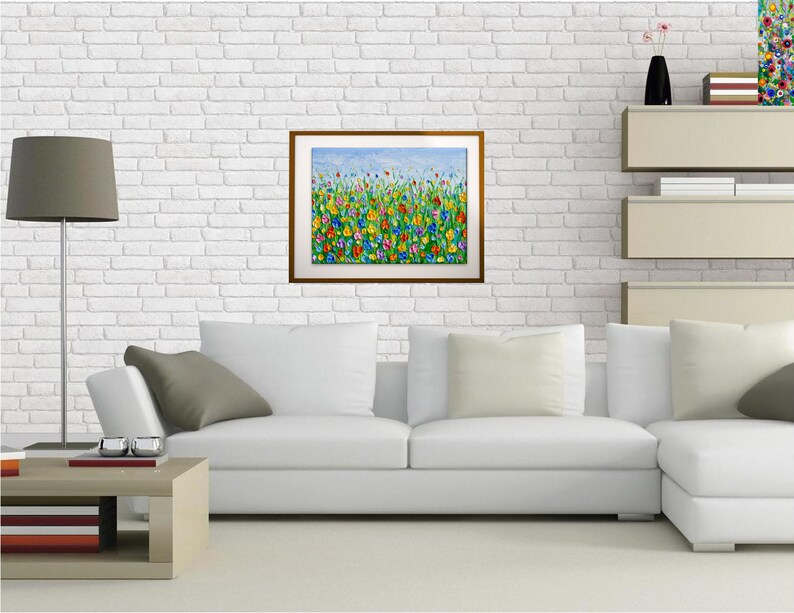 Puede incluir: Una pintura enmarcada que representa un campo vibrante de flores coloridas bajo un cielo azul. La obra de arte presenta una variedad de flores en tonos de rojo, amarillo, azul y verde. La pintura se exhibe en una pared de ladrillo blanco sobre un sof&aacute; blanco.