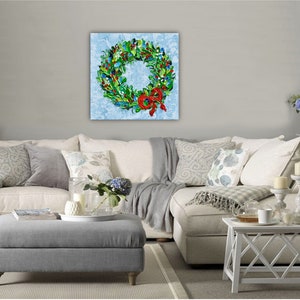 May include: A vibrant wreath painting on a light blue canvas. The wreath features green foliage, red berries, and a red bow. The artwork is displayed above a white sofa with grey and patterned cushions.
