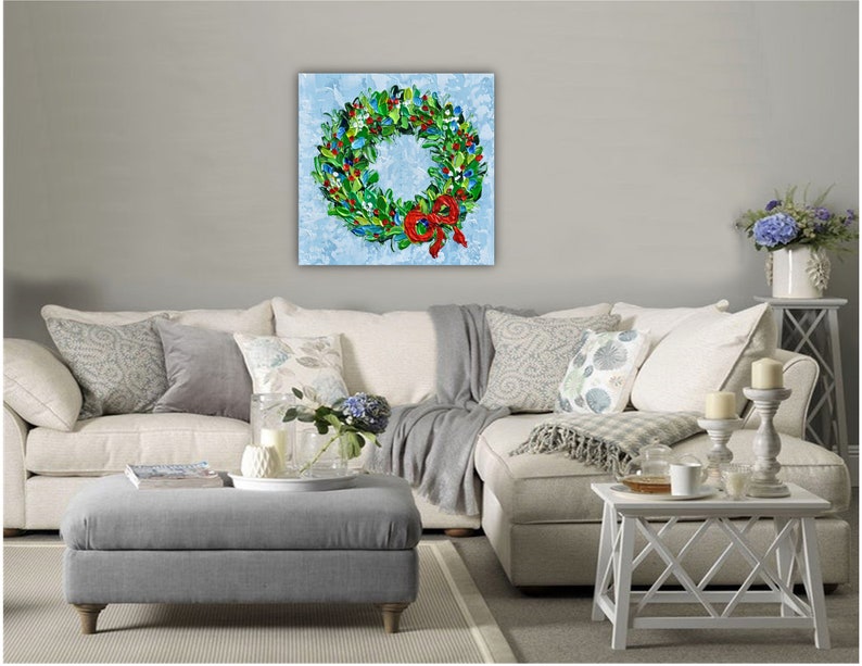 May include: A vibrant wreath painting on a light blue canvas. The wreath features green foliage, red berries, and a red bow. The artwork is displayed above a white sofa with grey and patterned cushions.