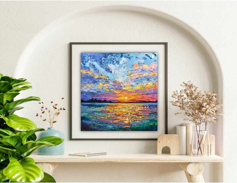 May include: Square framed painting of a sunset over water. The artwork features a vibrant sky with shades of blue, orange, and yellow. The water reflects the sunset's colors. The painting is displayed on a shelf with decorative items.