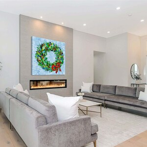 May include: A vibrant wreath painting with a red bow adorns a wall above a fireplace in a contemporary living room. The wreath showcases green foliage, blue details, and diverse colourful decorations. The room includes grey sofas, a white rug, and a marble coffee table.