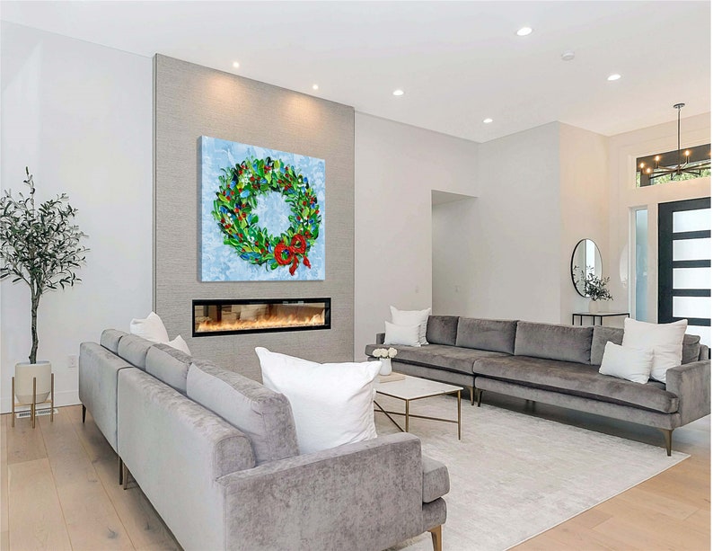 May include: A vibrant wreath painting with a red bow adorns a wall above a fireplace in a contemporary living room. The wreath showcases green foliage, blue details, and diverse colourful decorations. The room includes grey sofas, a white rug, and a marble coffee table.