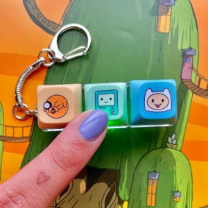 Puede incluir: Un llavero con tres colgantes cuadrados con diseños de personajes de dibujos animados. Los colgantes son de color beige, turquesa y azul, con imágenes de un perro, una consola de videojuegos y un personaje con un sombrero blanco. El llavero tiene un cierre plateado.