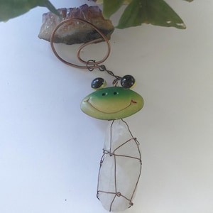 Könnte beinhalten: Ein skurriles Windspiel in Froschform. Der Frosch hat ein grün-gelbes Gesicht mit schwarzen Augen und einem lächelnden Mund. Ein klarer, länglicher Kristall ist darunter aufgehängt, umwickelt mit Kupferdraht. Das Windspiel ist an einer Kupferschlaufe befestigt.