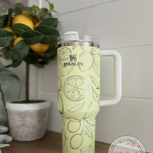 Pu&ograve; includere: Un bicchiere in acciaio inossidabile giallo con un coperchio e una maniglia bianchi. Il bicchiere ha un design a stampa di limone e il logo Stanley. Il bicchiere &egrave; appoggiato su una superficie di legno. Thistle Dew Lane &egrave; stampato su un adesivo sul bicchiere.