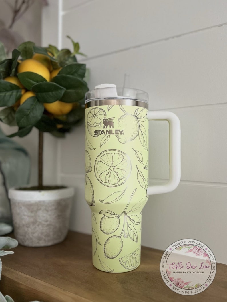 Pu&ograve; includere: Un bicchiere in acciaio inossidabile giallo con un coperchio e una maniglia bianchi. Il bicchiere ha un design a stampa di limone e il logo Stanley. Il bicchiere &egrave; appoggiato su una superficie di legno. Thistle Dew Lane &egrave; stampato su un adesivo sul bicchiere.