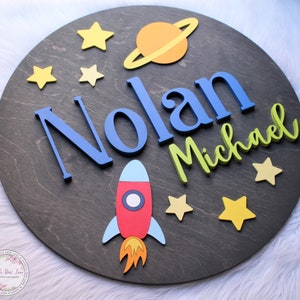 May include: A round wooden sign with a space theme. The sign features the names "Nolan Michael" in blue and green letters, a red rocket, a yellow and orange planet with rings, and yellow stars.