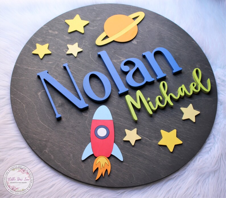 May include: A round wooden sign with a space theme. The sign features the names "Nolan Michael" in blue and green letters, a red rocket, a yellow and orange planet with rings, and yellow stars.
