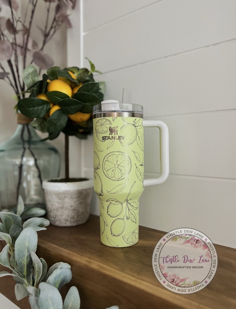 Pu&ograve; includere: Un bicchiere Stanley giallo e verde con un design a stampa di limone. Il bicchiere ha un coperchio bianco e una maniglia bianca. Il bicchiere &egrave; appoggiato su uno scaffale di legno con una pianta verde in un vaso.