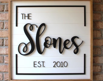 May include: A white wooden sign with a black frame and black lettering. The sign reads "The Stones Est. 2010".