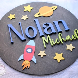 May include: A round wooden sign with a space theme. The sign features the names "Nolan Michael" in blue and green letters, a red rocket, a yellow and orange planet with rings, and yellow stars.