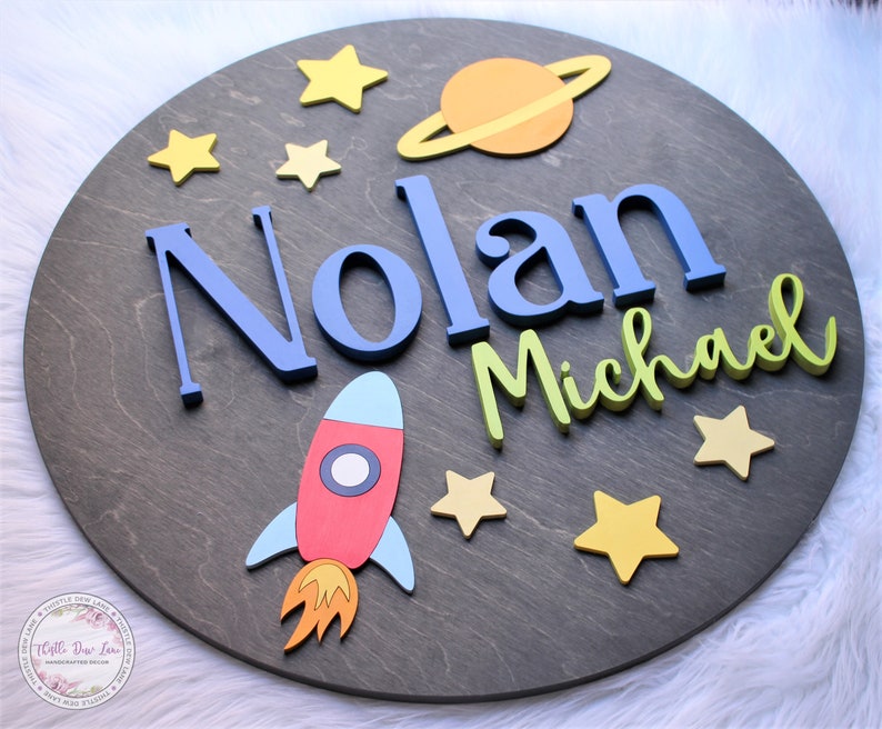 May include: A round wooden sign with a space theme. The sign features the names "Nolan Michael" in blue and green letters, a red rocket, a yellow and orange planet with rings, and yellow stars.