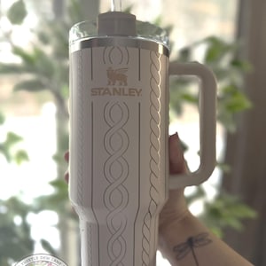May include: White stainless steel travel mug with a cable knit design and a gold Stanley logo. The mug has a black handle and a clear lid. The mug is being held by a person with a dragonfly tattoo on their wrist.