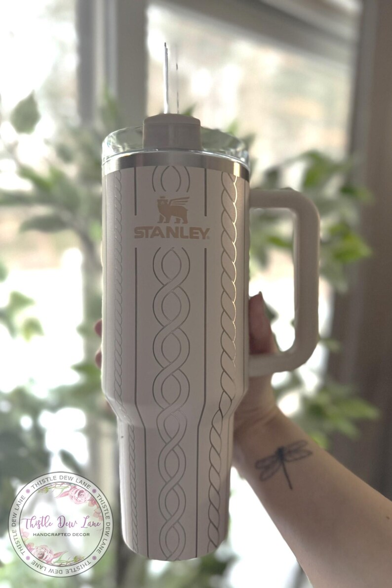 May include: White stainless steel travel mug with a cable knit design and a gold Stanley logo. The mug has a black handle and a clear lid. The mug is being held by a person with a dragonfly tattoo on their wrist.