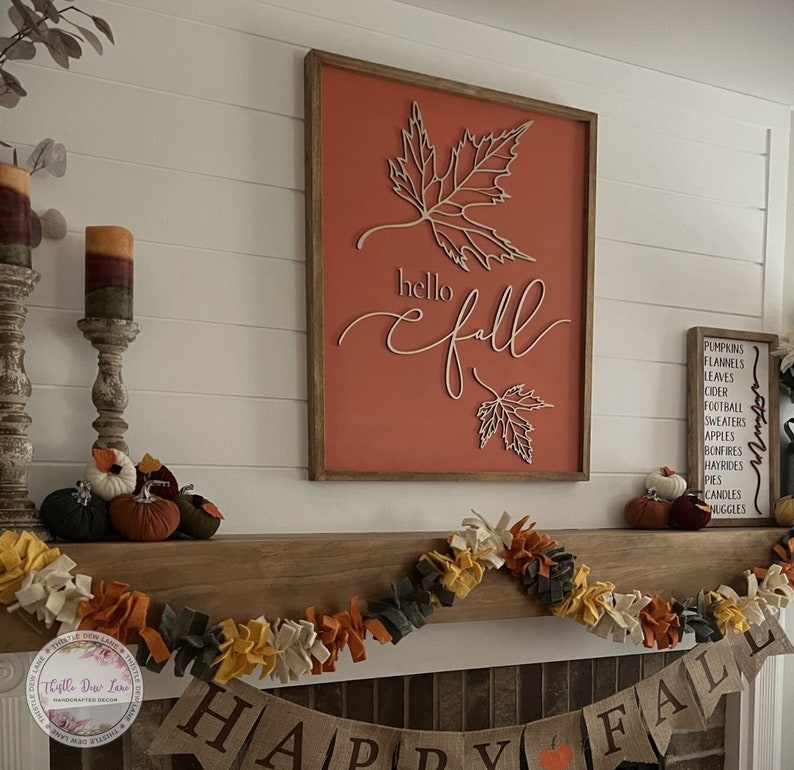 May include: A wooden sign with the words 'hello fall' in white script with a brown wood frame and a red background. The sign is decorated with two white paper cutouts of maple leaves. The sign is hanging on a white wall above a fireplace mantel. The mantel is decorated with a garland of brown, yellow, and gray felt balls. A banner with the words 'Happy Fall' is displayed on the mantel below the garland. The sign is a fall themed decoration.