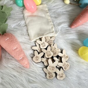 K&ouml;nnte beinhalten: Ein Satz aus Holz-Osterei-Such-Token in Form von Hasen mit verschiedenen Aktivit&auml;ten, die darauf geschrieben sind, wie "Filmabend", "Pizzaabend", "Zuhause bleiben", "Keine Hausarbeiten den ganzen Tag", "Neues Buch", "Kinder w&auml;hlen das Abendessen", "Basteln gehen", "Etwas pflanzen", "Drau&szlig;en spielen" und "Outdoor-Abenteurer-Jagd". Die Token befinden sich in einem wei&szlig;en Stoffbeutel und sind von pastellfarbenen Ostereiern umgeben.