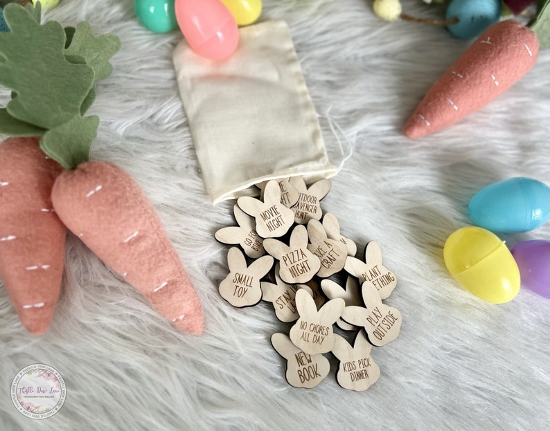K&ouml;nnte beinhalten: Ein Satz aus Holz-Osterei-Such-Token in Form von Hasen mit verschiedenen Aktivit&auml;ten, die darauf geschrieben sind, wie "Filmabend", "Pizzaabend", "Zuhause bleiben", "Keine Hausarbeiten den ganzen Tag", "Neues Buch", "Kinder w&auml;hlen das Abendessen", "Basteln gehen", "Etwas pflanzen", "Drau&szlig;en spielen" und "Outdoor-Abenteurer-Jagd". Die Token befinden sich in einem wei&szlig;en Stoffbeutel und sind von pastellfarbenen Ostereiern umgeben.